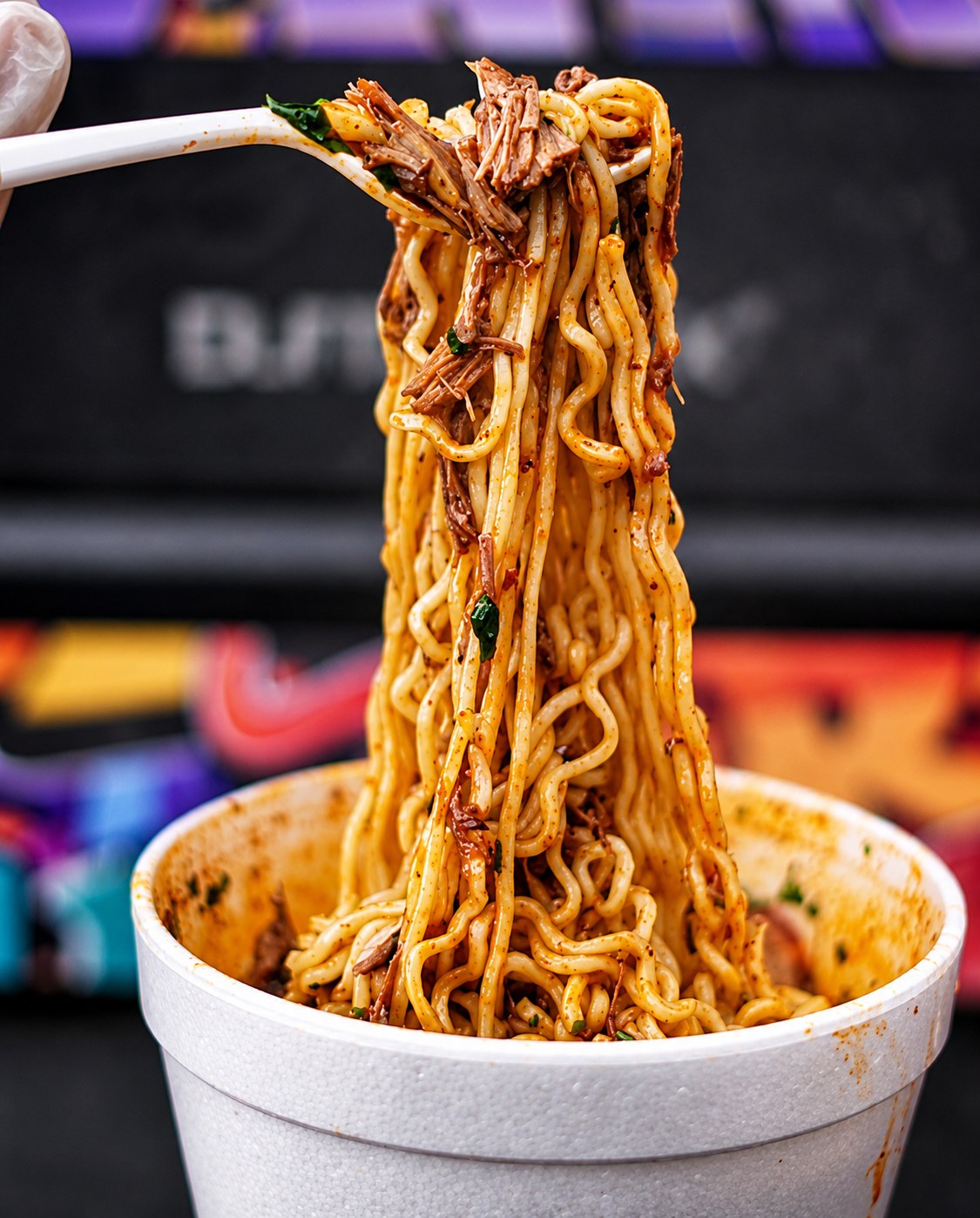 Birria ramen noodles lifted high out of a styrofoam cup of consom&eacute;