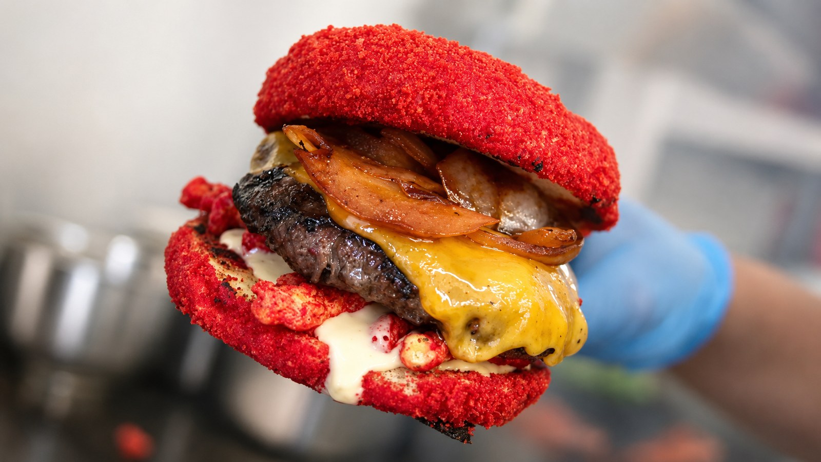 A Hot Cheeto Burger held in a blue-gloved hand, cheese and white sauce dripping from the red crusted bun