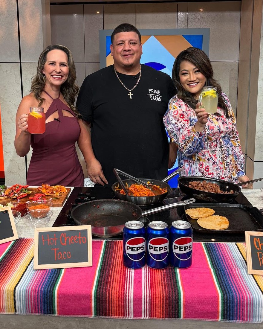 Anthony Solis in a Prime Tacos tee cooking on a TV studio set with two hosts