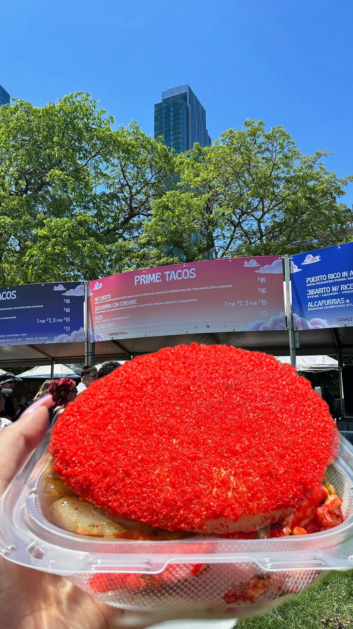 A Hot Cheeto Burger held up in a clamshell in front of the Prime Tacos menu board at the festival stand in Grant Park